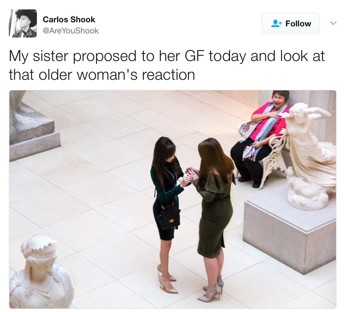 Tweet from Carlos Shook: 'My sister proposed to her GF today and look at that older woman's reaction'. Image: Two women stand inside what appears to be a museum, surrounded by sculptures. As one woman proposes to the other, placing a ring on the other woman's finger, an older woman who is sitting nearby looks on with sheer joy.